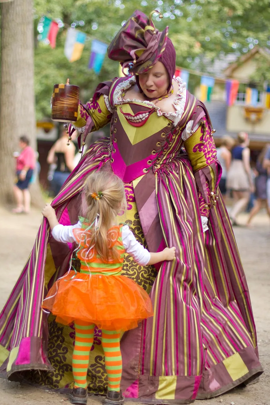 A 16th-century court jester in a brown, lavender, green and fuchsia striped satin motley. A little girl dressed as a fairy in orange and green, with tiny orange wings, examines the jester's motley.