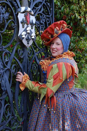 16th-century court jester in a blue-and-red brocade gown with green Italianate sleeves, with orange and green silk dagges, a goldenrod shift with redwork embroidery, a blue pudding cap with gold embroidery, and a brick red spotted riding cap with a multicolor coxcomb crown, at a gate at Muncaster Castle in Cumbria, UK.