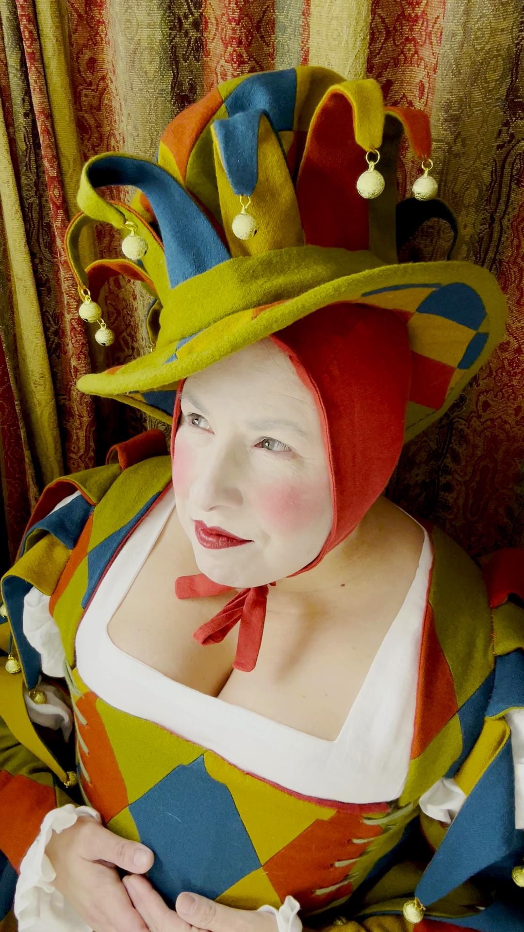 16th-century court jester in a patchwork motley in green, russet, blue and gold wool, with a linen shift and a red linen pudding-cap.